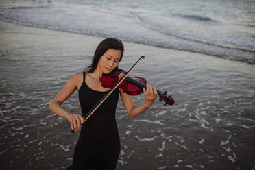 Woman playing violin in front of sea at beach - GMLF01229