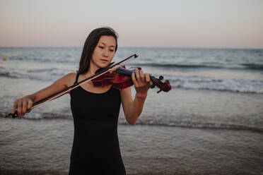 Woman playing violin in front of sea at sunset - GMLF01230