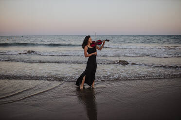 Woman playing violin standing in front of sea at sunset - GMLF01234