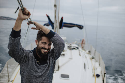Lächelnder junger Mann hält das Seil eines Segelboots im Mittelmeer - GMLF01238