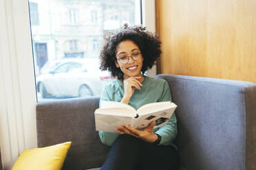Smiling woman reading book sitting on sofa - OYF00657