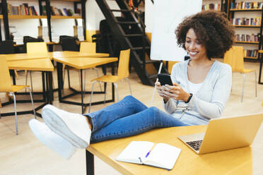 Lächelnde Geschäftsfrau, die am Schreibtisch in einem kreativen Büro eine SMS auf ihrem Smartphone schreibt - OYF00681