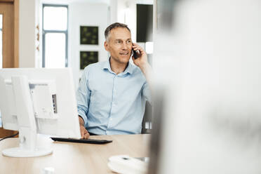 Mature businessman talking on smart phone sitting at computer at desk in office - JOSEF06470