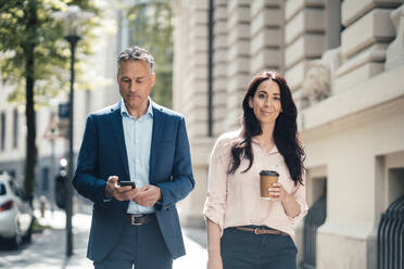 Geschäftsfrau mit Einweg-Kaffeebecher, die an einem Geschäftsmann vorbeigeht, der auf dem Gehweg ein Smartphone benutzt - JOSEF06479