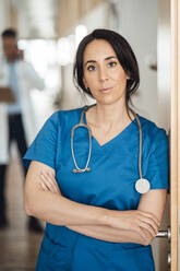 Confident doctor with arms crossed leaning on door in hospital - JOSEF06482