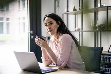 Lächelnde Geschäftsfrau mit Laptop und Kaffee am Schreibtisch in einem Coworking-Büro - JOSEF06497