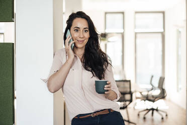 Geschäftsfrau mit Kaffeetasse, die in einem Coworking-Büro mit ihrem Smartphone telefoniert - JOSEF06504