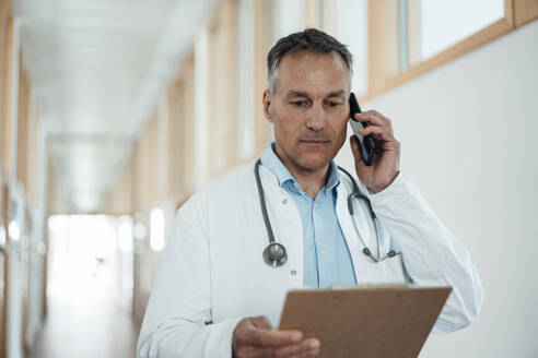 Älterer Arzt, der die Krankenakte prüft und mit einem Smartphone im Krankenhaus telefoniert - JOSEF06521
