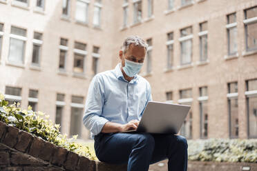 Businessman in protective face mask using laptop at office park - JOSEF06522