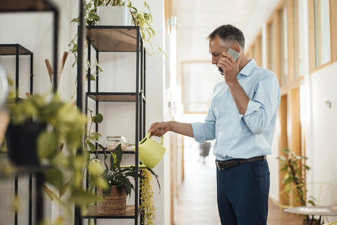 Businessman talking on mobile phone watering plant at corridor in office - JOSEF06528