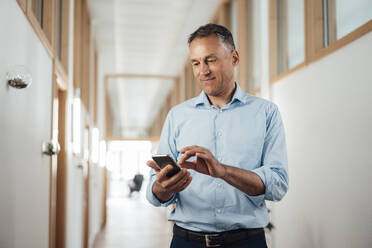 Älterer Geschäftsmann, der sein Smartphone im Korridor im Büro benutzt - JOSEF06530