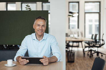 Smiling mature businessman with tablet PC sitting at desk in coworking office - JOSEF06531