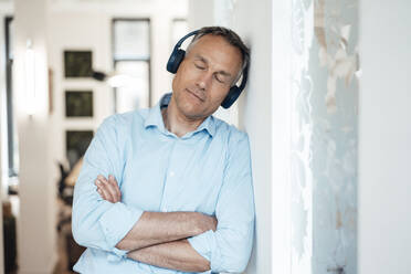 Businessman with eyes closed listening music and leaning on wall in office - JOSEF06533