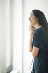 Happy businesswoman talking on telephone by window in coworking office - JOSEF06596