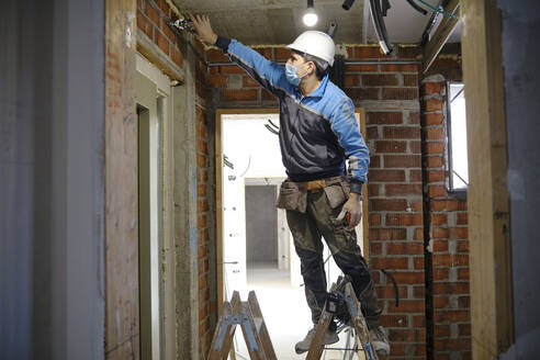 Electrician working with tool standing on ladder at construction site - AGOF00214