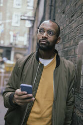 Nachdenklicher Mann mit Smartphone an der Wand lehnend - AMWF00085