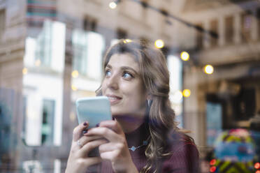 Nachdenkliche Frau mit Smartphone durch das Fenster eines Cafés gesehen - AMWF00088