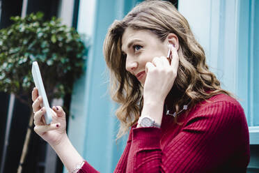 Lächelnde Frau mit Smartphone, die Bluetooth-Kopfhörer einstellt - AMWF00097