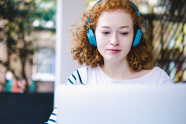 Redhead woman using laptop and listening music on headphones in cafe - AMWF00118