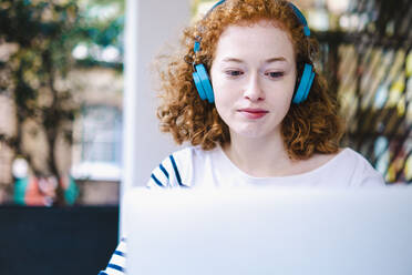 Rothaarige Frau benutzt Laptop und hört Musik über Kopfhörer in einem Cafe - AMWF00118