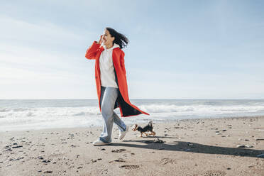 Lächelnde Frau, die mit ihrem Yorkshire-Terrier am Strand spazieren geht, an einem sonnigen Tag - OMIF00438