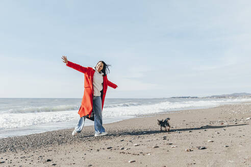 Fröhliche Frau mit ausgestreckten Armen bei einem Hund am Strand - OMIF00443