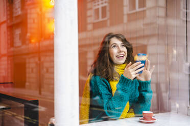 Fröhliche Frau mit Mobiltelefon durch das Glas eines Cafés gesehen - AMWF00140