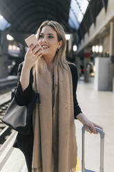 Smiling woman with luggage talking on speaker phone at station - JRVF02625