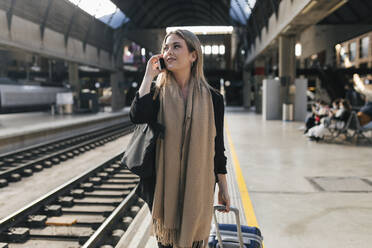 Frau mit Gepäck auf Rädern, die am Bahnsteig mit einem Mobiltelefon spricht - JRVF02626