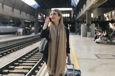 Woman with wheeled luggage talking on mobile phone at platform - JRVF02626