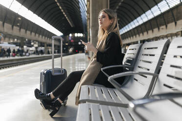 Nachdenkliche Frau mit Smartphone und Gepäck auf Rädern am Bahnhof - JRVF02627