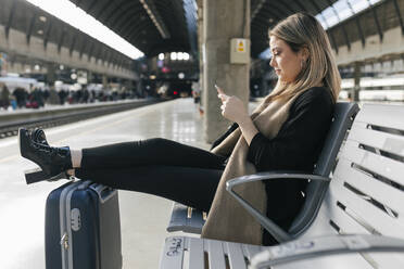 Woman using smart phone and leaning legs on wheeled luggage at station - JRVF02628