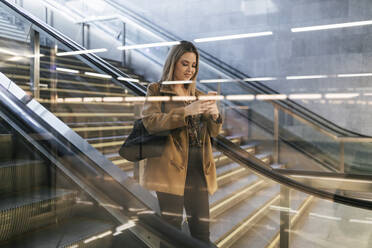 Lächelnde Frau, die ein Smartphone auf einer Rolltreppe in einer U-Bahn-Station benutzt - JRVF02668