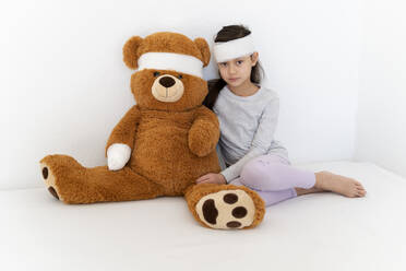 Bandage on girl with teddy bear sitting in front of white wall - DRF01803