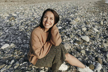 Fröhliche junge Frau sitzt auf Steinen am Strand - OMIF00456