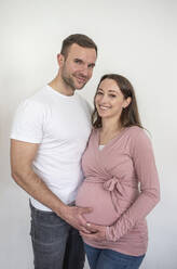Lächelnder Mann und Frau, die ihren schwangeren Bauch vor einer weißen Wand berühren - BFRF02401