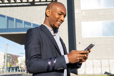 Side view of positive bald African American male in formal clothes browsing on smartphone while standing near building on sunny street of city - ADSF33370