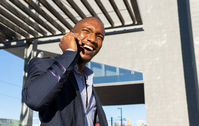 Positive bald African American male in formal clothes having phone conversation while standing near building on sunny street of city - ADSF33373