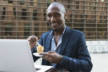 Positiver afroamerikanischer männlicher Unternehmer mit einer Tasse Kaffee, der in die Kamera schaut, am Tisch mit Laptop auf der Terrasse eines Cafés - ADSF33380