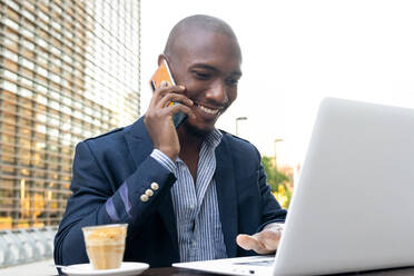 Fröhlicher afroamerikanischer männlicher Unternehmer im Anzug, der ein Telefongespräch führt, während er an einem Tisch mit Kaffee auf der Terrasse eines Cafés auf seinem Laptop surft - ADSF33381