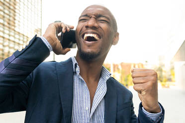Fröhlicher afroamerikanischer männlicher Unternehmer im Anzug führt ein Telefongespräch am Tisch mit Kaffee auf der Terrasse eines Cafés - ADSF33383