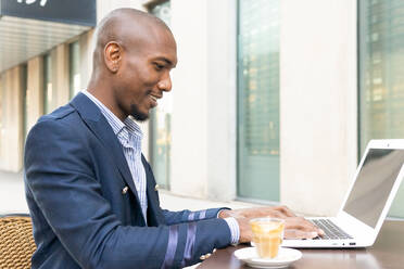 Seitenansicht eines positiven afroamerikanischen männlichen Unternehmers, der mit einem Laptop auf der Terrasse und einer Tasse Kaffee in einem Café am Tisch sitzt - ADSF33386