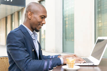 Side view of positive African American male entrepreneur sitting at table with laptop on terrace and cup of coffee in cafe - ADSF33386