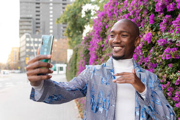 Lächelnder afroamerikanischer Mann in stilvollem Outfit, der mit einem modernen Smartphone Selbstporträt führt, während er in der Nähe blühender Büsche auf der Straße steht - ADSF33389
