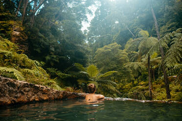 Rückenansicht eines jungen Mannes ohne Hemd, der im klaren Wasser eines Pools in einem exotischen Wald mit grünen Palmen auf den Azoren, Sao Miguel, schwimmt - ADSF33398