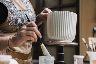 Crop anonymous female potter in apron painting handmade clay pot in professional workshop on blurred background - ADSF33400