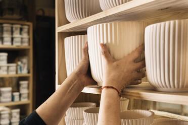 Crop anonymous female with bracelet standing near wooden shelves while arranging white clay pots in workshop - ADSF33402