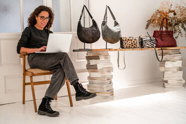 Ganzkörperaufnahme einer Designerin, die auf einem modernen Netbook surft, während sie in der Nähe eines Tisches mit verschiedenen modischen Handtaschen in einem Ausstellungsraum für Lichtdesign sitzt - ADSF33413