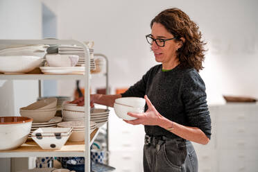 Seitenansicht einer Designerin mit Brille und Schalen, die in der Nähe von Regalen mit einer Sammlung von kreativem Geschirr in einer hellen Werkstatt steht - ADSF33414