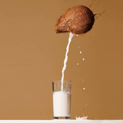 Milk pouring from fresh whole coconut into glass served on table against brown background - ADSF33417